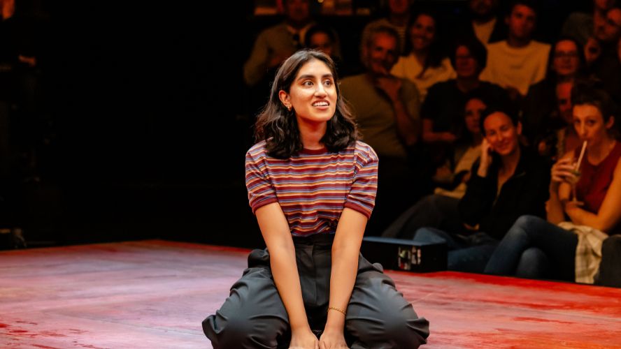 Ambika mod kneels on stage @sohoplace wearing a striped tshirt and black trousers