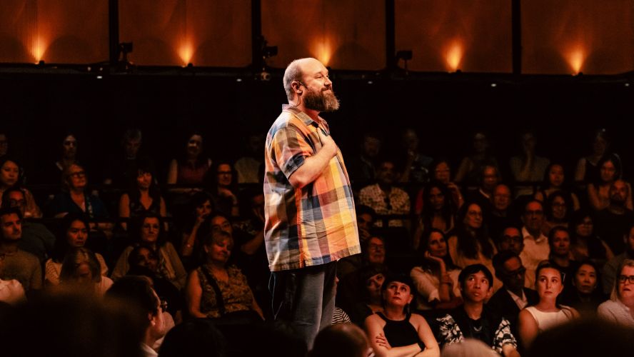 Jonny Donahoe stands on stage @sohoplace with his hand on his chest. He is wearing a colourful check shirts and jeans.