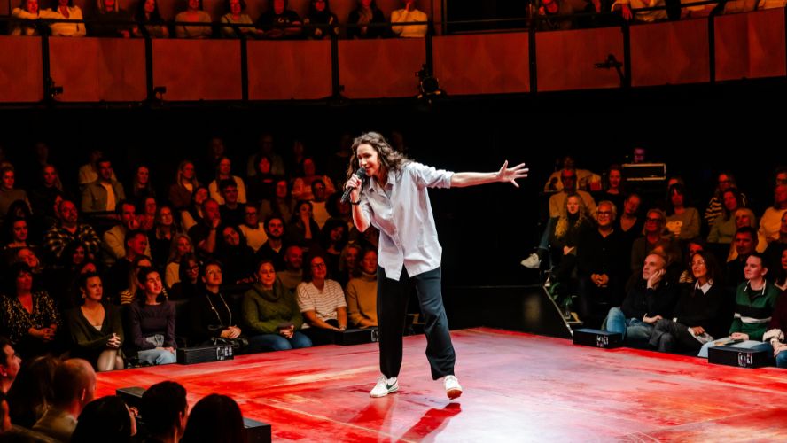 Minnie Driver wears a blue shirt and black trousers, holding a microphone while talking to the audience