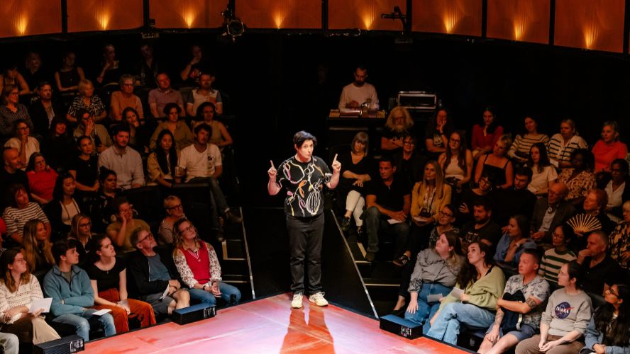 Sue Perkins stands on stage holding her hands up, wearing w black shirt with colourful patterns