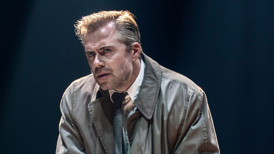 Rory Keenan plays Alec Leamas. He's looking confused and wearing a trench coat, shirt and tie