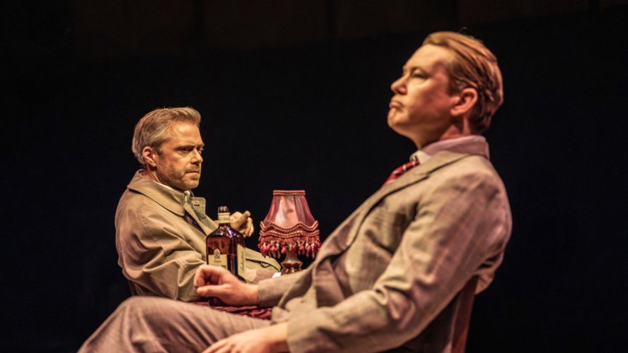 Rory Keenan plays Alec Lemas, sitting opposite Mat Betteridge who plays Karl Riemeck/Kiever. There is a lamp and a bottle of alcohol on the table between them
